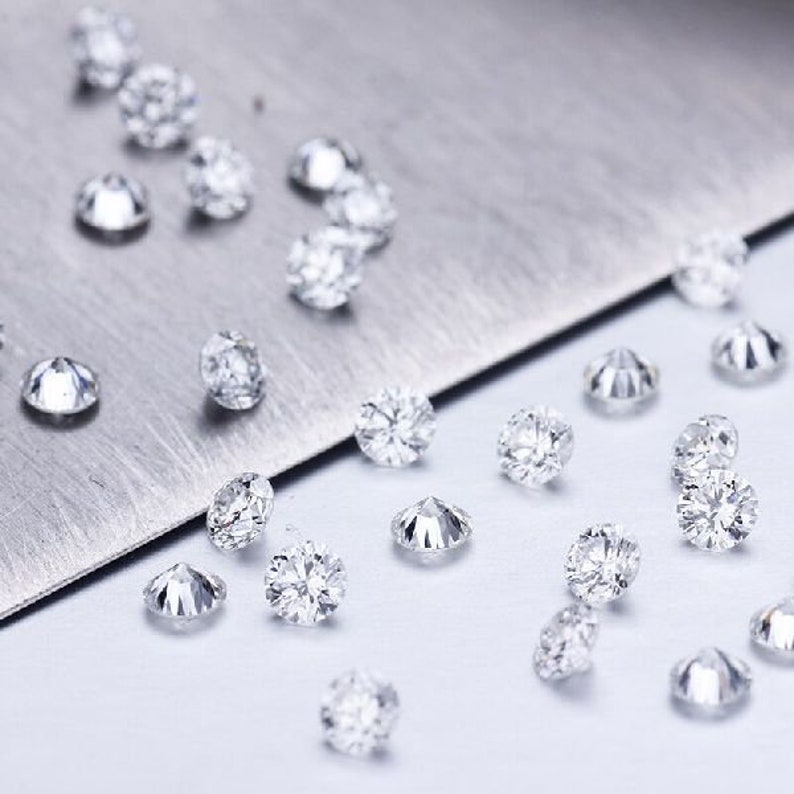 Puede incluir: Una colecci&oacute;n de peque&ntilde;os diamantes redondos y transparentes esparcidos sobre una superficie blanca. Los diamantes son de varios tama&ntilde;os y est&aacute;n dispuestos en un patr&oacute;n aleatorio.