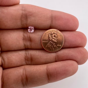 May include: A pink gemstone, approximately the size of a US penny, is shown in a hand. The gemstone is a square cushion cut with facets.  "In God We Trust" and "Liberty" are visible on the penny.
