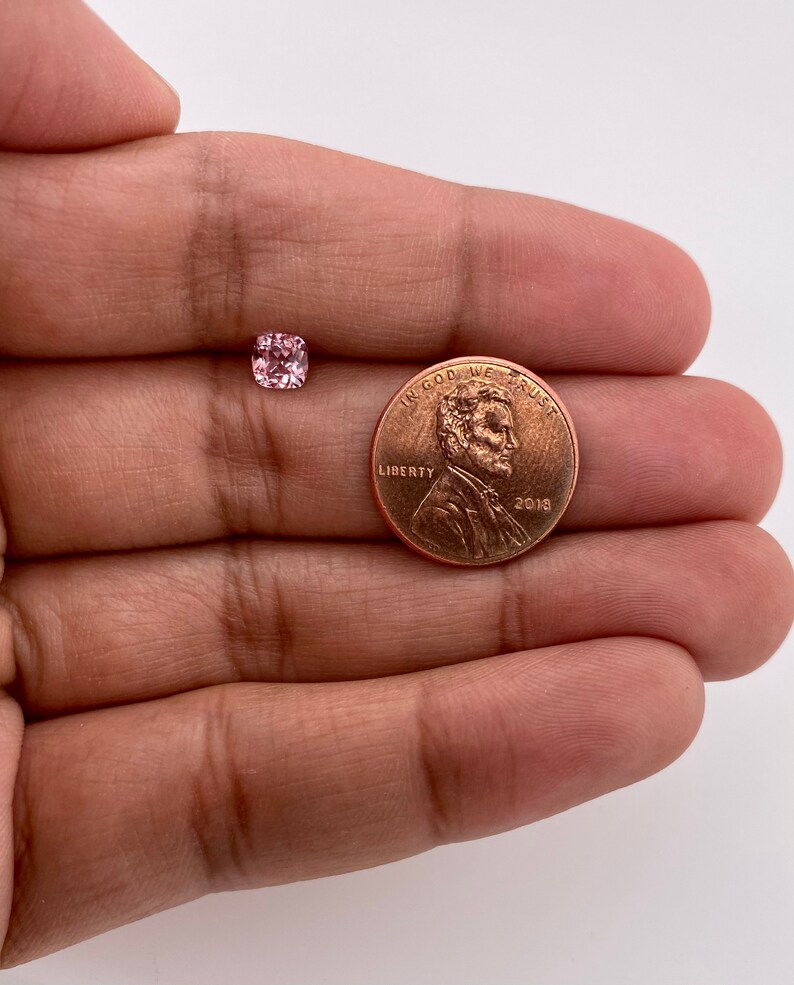 May include: A pink gemstone, approximately the size of a US penny, is shown in a hand. The gemstone is a square cushion cut with facets.  "In God We Trust" and "Liberty" are visible on the penny.