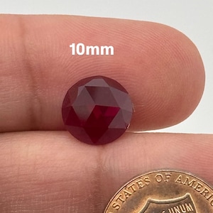 May include: A deep red, faceted gemstone measuring 10mm in diameter. The gemstone is held in a hand, with a US penny in the background for size comparison.