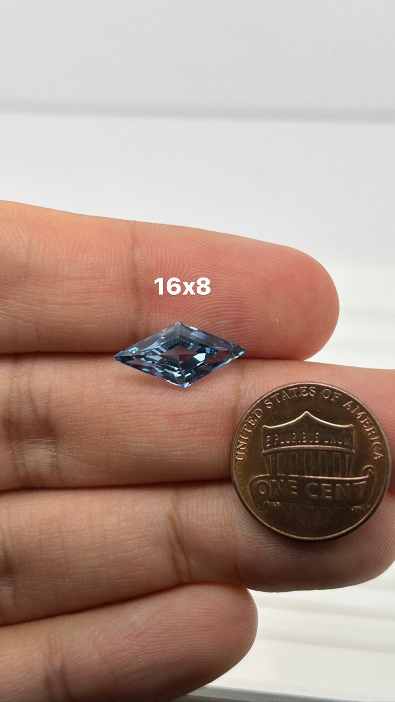 Peut inclure: Une pierre pr&eacute;cieuse bleu clair en forme de cerf-volant, mesurant 16x8, est tenue dans une main &agrave; c&ocirc;t&eacute; d'un penny am&eacute;ricain pour comparaison de taille.