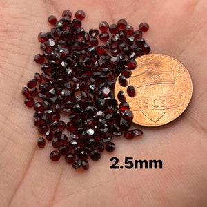 May include: A pile of small, dark red, round gemstones, approximately 2.5 mm in diameter, next to a US penny for size comparison.