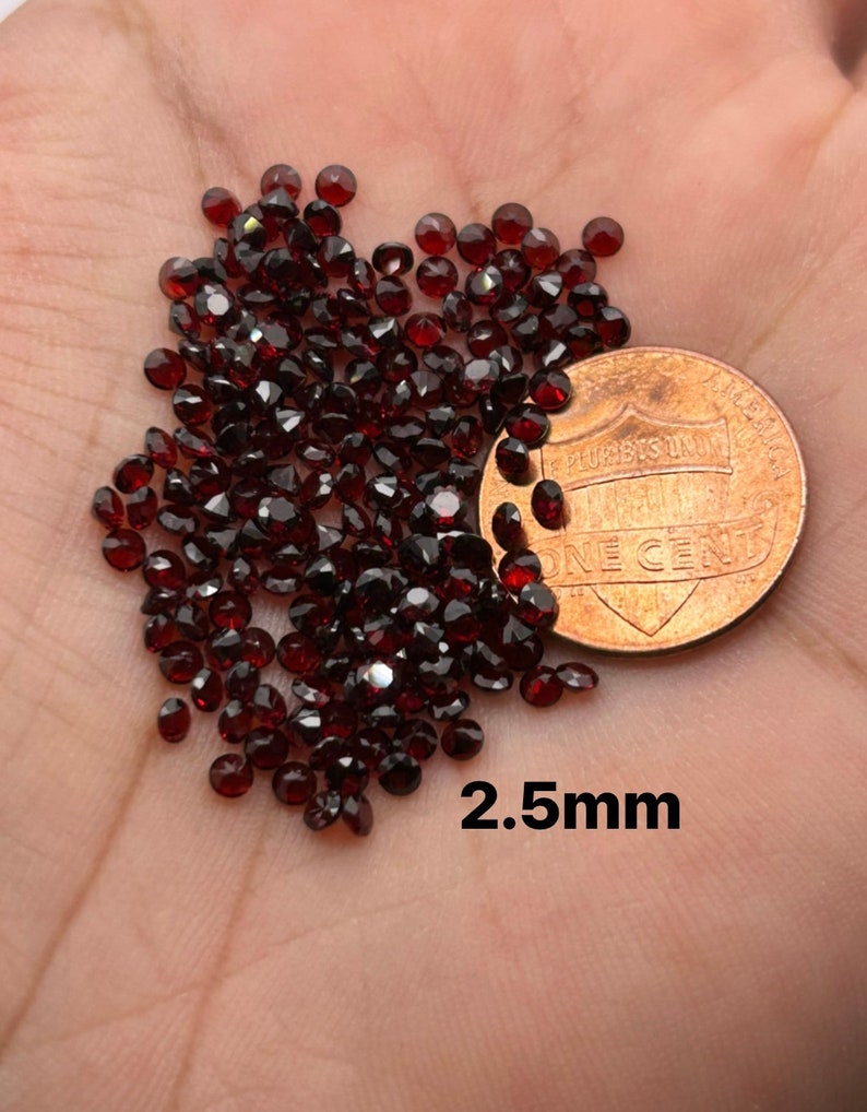 May include: A pile of small, dark red, round gemstones, approximately 2.5 mm in diameter, next to a US penny for size comparison.