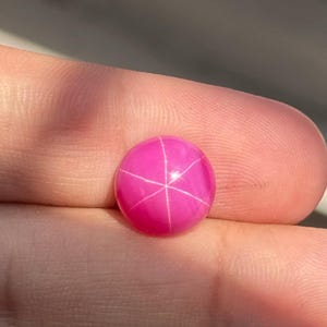 May include: A round, vibrant hot pink cabochon gemstone featuring a distinct six-rayed star pattern across its smooth, polished surface. The stone displays a bright, saturated magenta hue with a sharp, centered asterism effect visible on top.