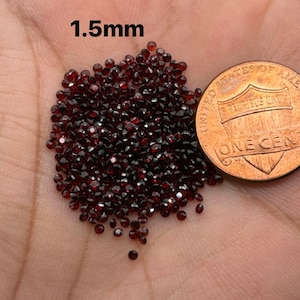 May include: A pile of small, dark red, faceted gemstones, approximately 1.5mm in size, next to a US penny for scale.