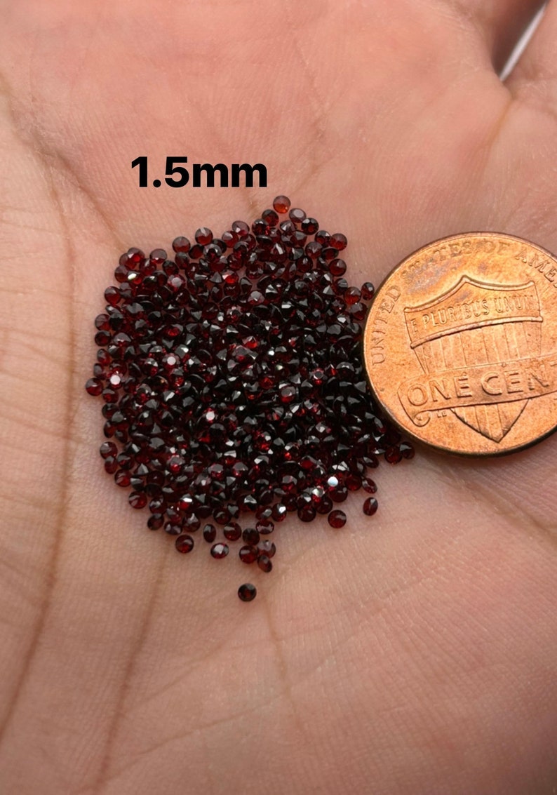 May include: A pile of small, dark red, faceted gemstones, approximately 1.5mm in size, next to a US penny for scale.