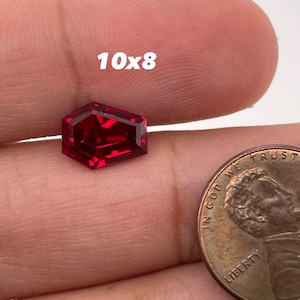 May include: A deep red gemstone with a faceted, octagonal shape. The gemstone is held in a hand, with a penny for scale. The text "10x8" is visible in the image.