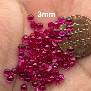 May include: A pile of 3mm round ruby red cabochon gemstones, next to a US penny for size comparison.