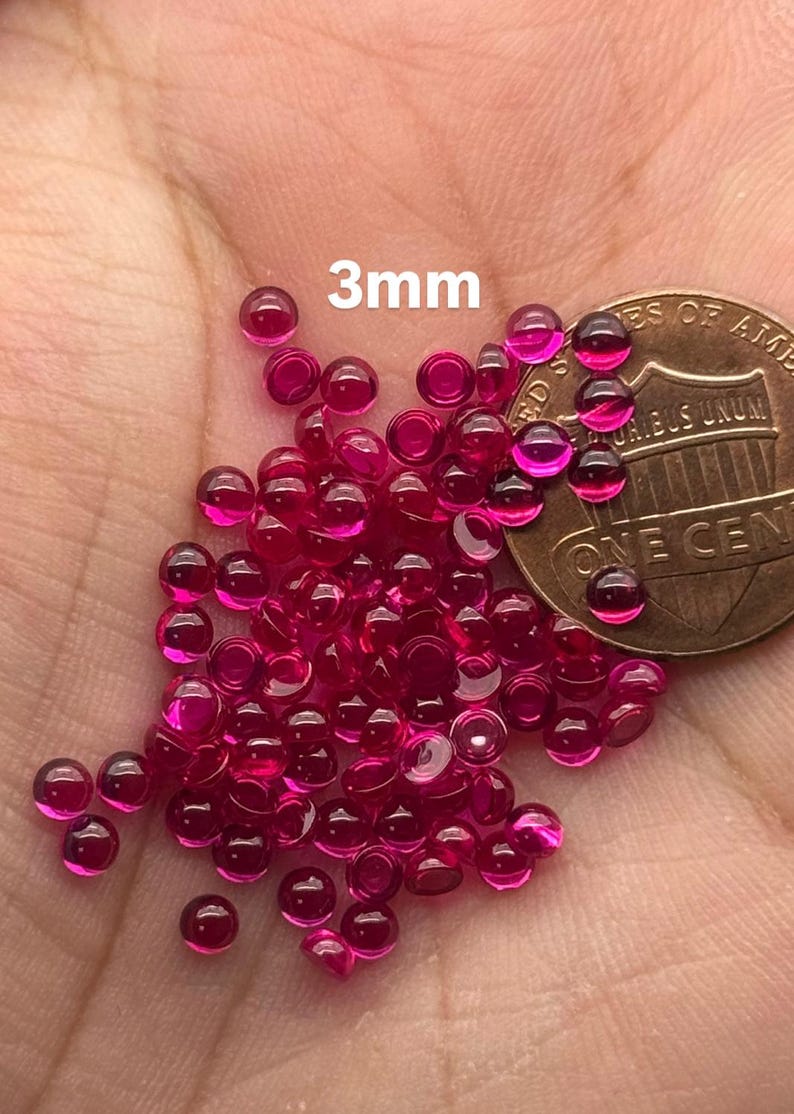 May include: A pile of 3mm round ruby red cabochon gemstones, next to a US penny for size comparison.
