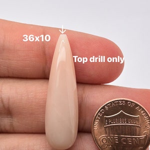 May include: A pale pink teardrop-shaped cabochon with a smooth, polished surface. The cabochon is approximately 36mm x 10mm and is held in a hand for size comparison. The text "Top drill only" is written on the image.