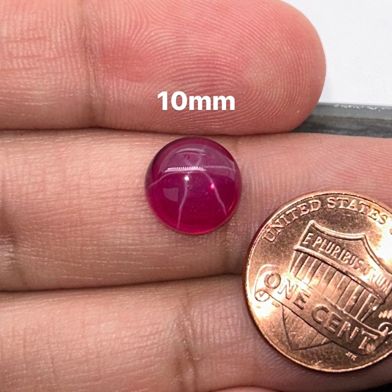 May include: A 10mm round, pink star ruby cabochon gemstone with white star-like inclusions. The gemstone is shown next to a US penny for size comparison.