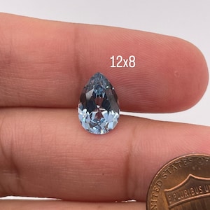 May include: A light blue pear-shaped gemstone, measuring 12x8, is held in a hand for size comparison. A US penny is also shown in the image.