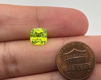 May include: A bright green gemstone with a cushion cut, displayed on a hand next to a US penny for size comparison.
