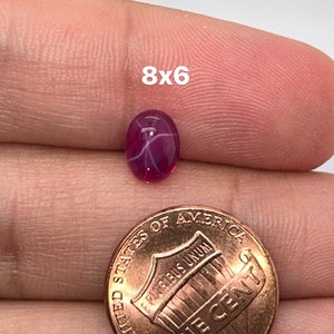 May include: A pink star ruby gemstone with a 8x6 mm oval shape. The gemstone is held in a hand with a US penny for size comparison.