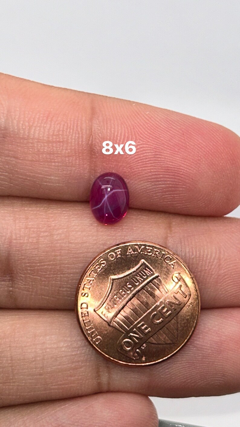 May include: A pink star ruby gemstone with a 8x6 mm oval shape. The gemstone is held in a hand with a US penny for size comparison.