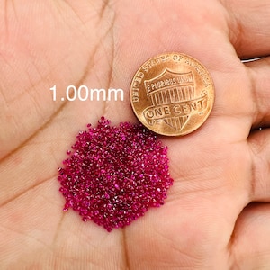 May include: A pile of small, red ruby gemstones, approximately 1.00mm in size, next to a US penny for scale.