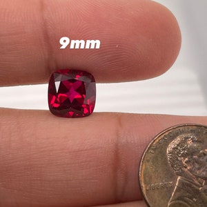 May include: A vibrant, cushion-cut, red gemstone, approximately 9mm in size, is displayed. The gemstone is held near a penny for size comparison. The gem has a star-like pattern within its facets.