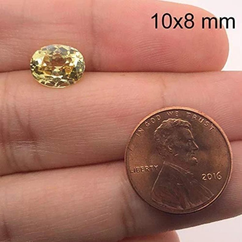 May include: A yellow oval gemstone with a faceted cut, measuring 10x8 millimetres. The gemstone is held in a hand, with a US penny in the background for size comparison.
