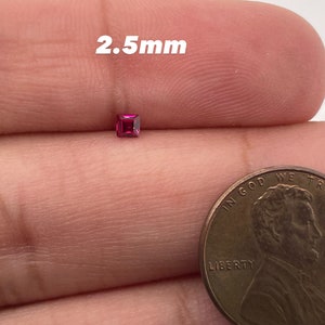 May include: A small, square, pink gemstone measuring 2.5 mm in size. The gemstone is held in a person's hand next to a US penny for size comparison.