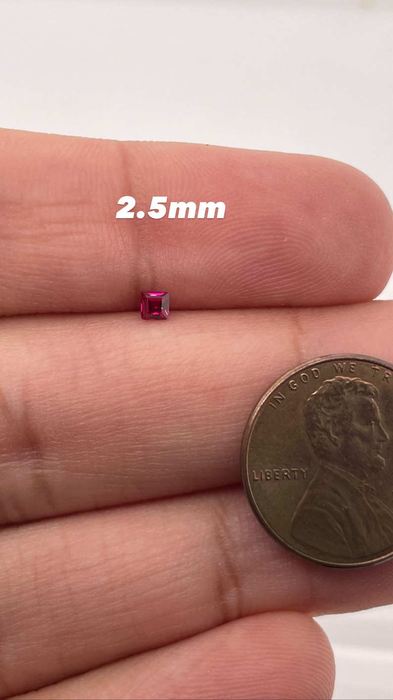 May include: A small, square, pink gemstone measuring 2.5 mm in size. The gemstone is held in a person's hand next to a US penny for size comparison.