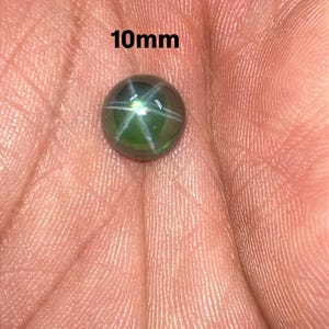 May include: A 10mm green star sapphire gemstone. The gemstone has a six-rayed star pattern and is set against a hand. The text "10mm" is visible in the image.