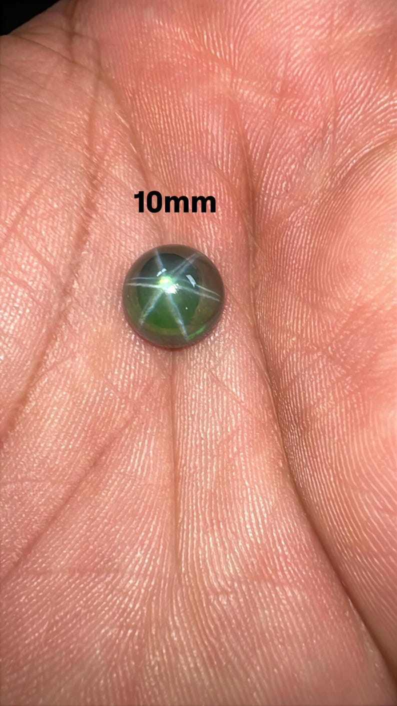 May include: A 10mm green star sapphire gemstone. The gemstone has a six-rayed star pattern and is set against a hand. The text "10mm" is visible in the image.