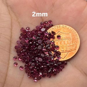 May include: A collection of small, round, dark pink gemstones, approximately 2mm in diameter, are scattered across a hand. A U.S. penny is also present, providing a size reference. The text "2mm" is visible.