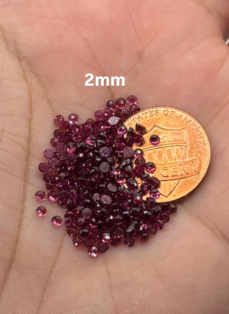 May include: A collection of small, round, dark pink gemstones, approximately 2mm in diameter, are scattered across a hand. A U.S. penny is also present, providing a size reference. The text "2mm" is visible.