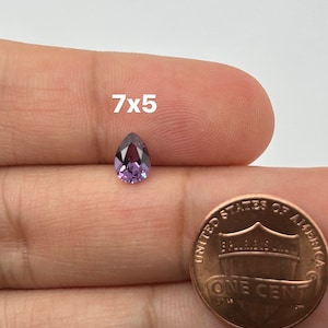 May include: A pear-shaped purple gemstone, measuring 7x5 mm, is held in a hand next to a US penny for size comparison.