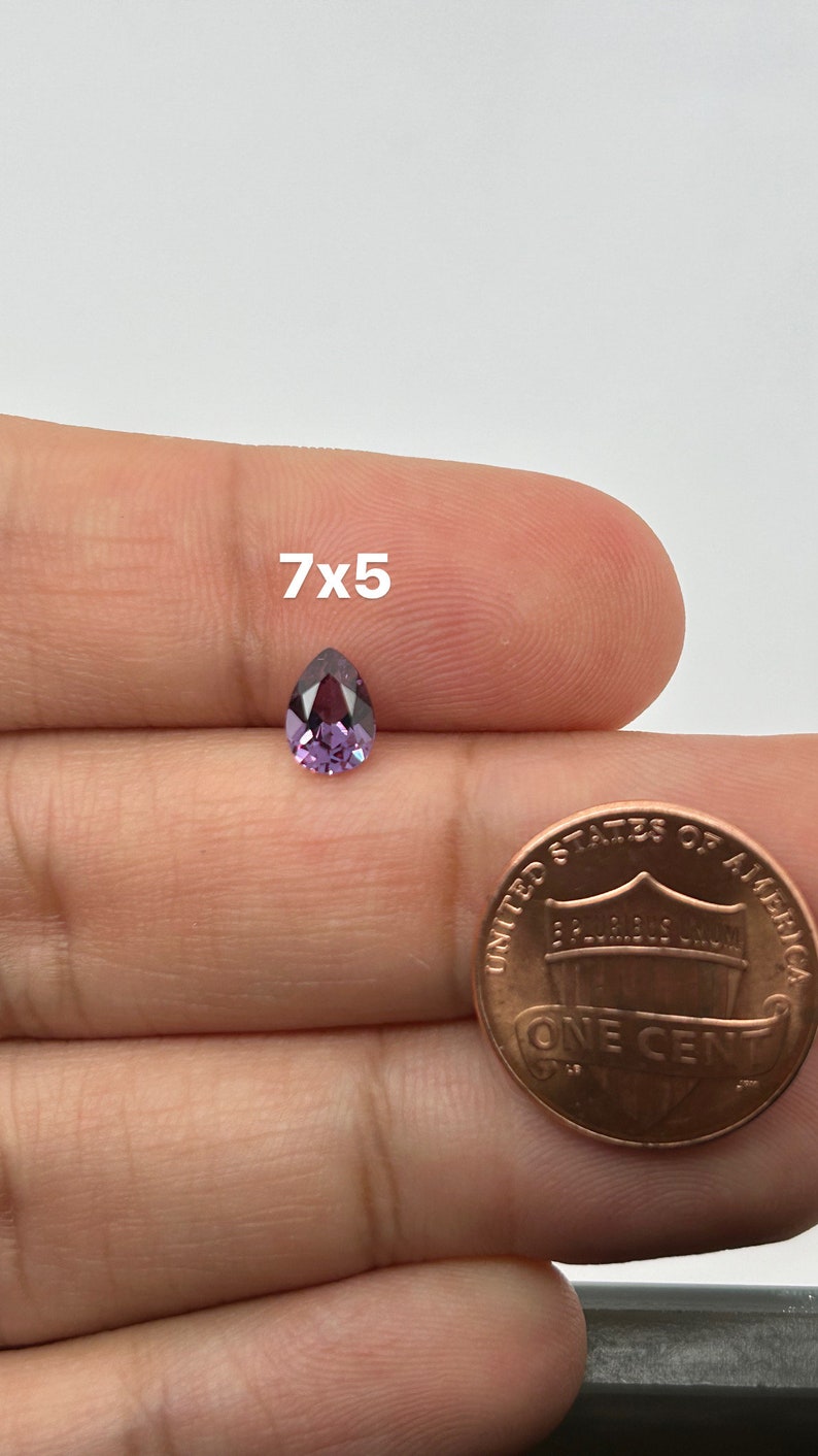May include: A pear-shaped purple gemstone, measuring 7x5 mm, is held in a hand next to a US penny for size comparison.
