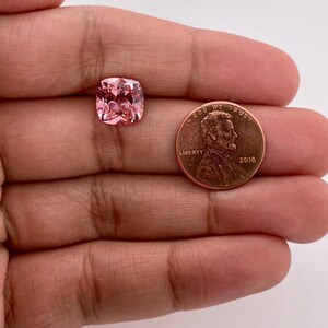 May include: A pink cushion-cut gemstone, approximately the size of a US penny, with a faceted surface.