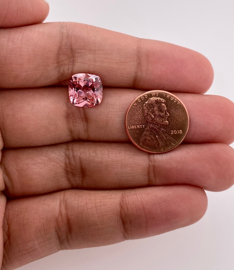 May include: A pink cushion-cut gemstone, approximately the size of a US penny, with a faceted surface.