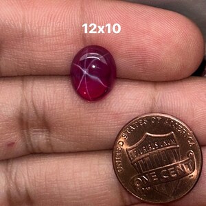May include: A 12x10 mm oval-shaped, pink ruby gemstone with a star-like pattern inside. The ruby is being held in a hand, next to a US penny for size comparison.
