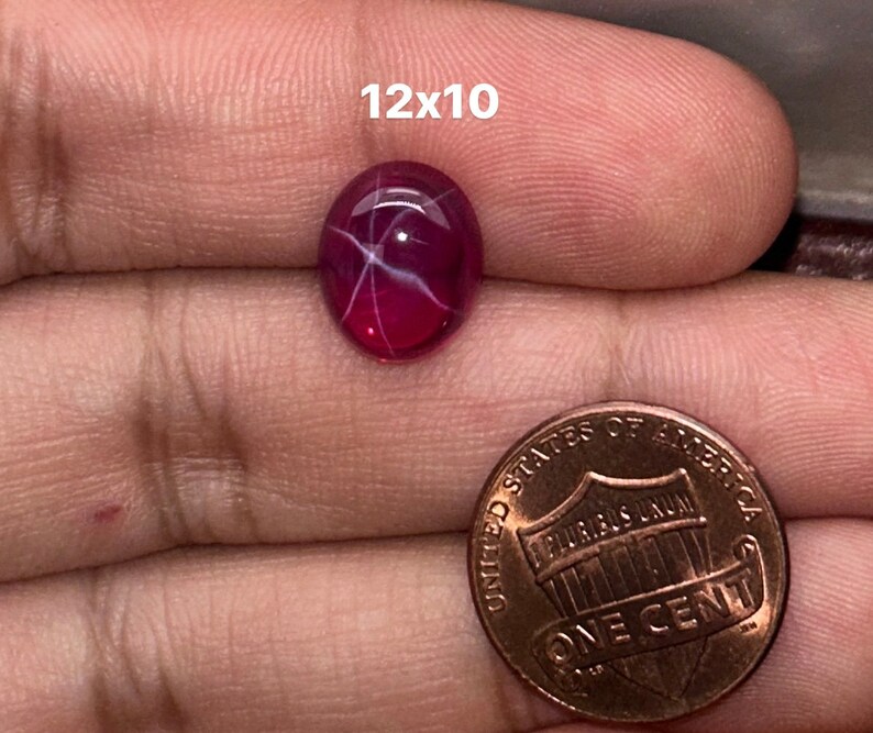 May include: A 12x10 mm oval-shaped, pink ruby gemstone with a star-like pattern inside. The ruby is being held in a hand, next to a US penny for size comparison.