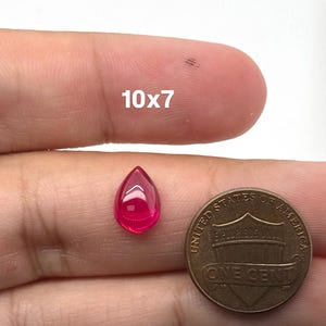 May include: A pear-shaped, translucent, vibrant pink gemstone measuring 10x7 millimeters, positioned next to a United States one-cent copper coin for scale. The gemstone features a smooth, polished surface with a distinct teardrop silhouette.