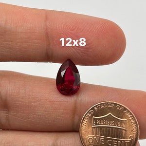 May include: A deep red pear-shaped gemstone, approximately 12x8 mm, held in a hand next to a US penny for size comparison.