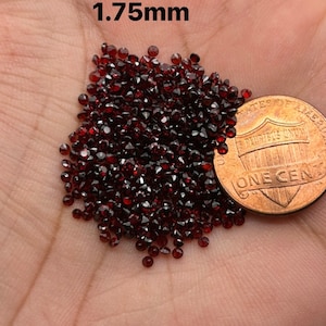 May include: A pile of small, dark red, round gemstones, approximately 1.75mm in diameter, next to a US penny for scale.
