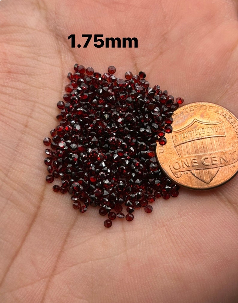 May include: A pile of small, dark red, round gemstones, approximately 1.75mm in diameter, next to a US penny for scale.