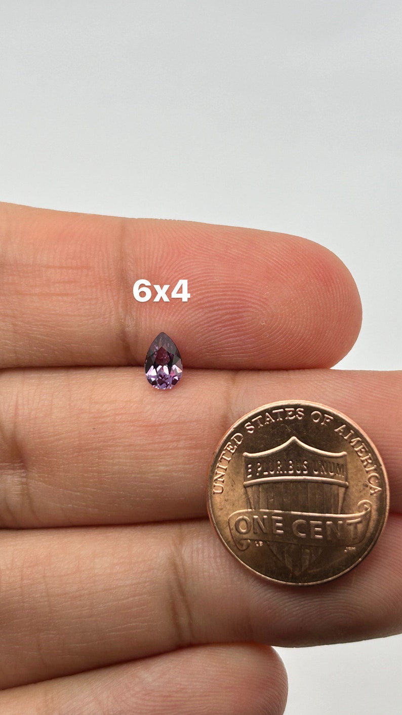 May include: A pear-shaped purple gemstone measuring 6x4 millimeters, shown next to a US penny for size comparison.