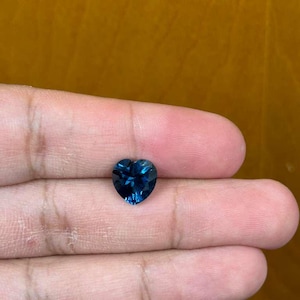 May include: A heart-shaped, deep blue gemstone. The gem is held in a hand, showcasing its facets and color. The stone's shape and color suggest it could be a sapphire or similar precious stone, ideal for jewelry.