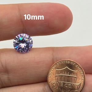 May include: A round, faceted, light purple gemstone, approximately 10mm in diameter, is held between fingers. A United States one-cent coin is next to the gemstone. The text "10mm" is above the gemstone.