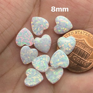 May include: A group of 8mm white opal heart-shaped cabochons, some with iridescent rainbow flashes, next to a UK penny for size comparison.