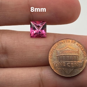 May include: A pink square-shaped gemstone measuring 8mm in size. The gemstone is held in a hand for size comparison next to a US penny.
