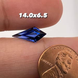 May include: A deep blue gemstone with a kite shape, measuring 14.0 x 6.5 mm, is held in a hand next to a UK penny for size comparison.
