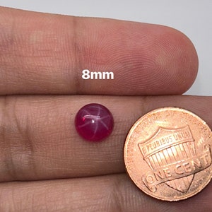 May include: A pink star ruby gemstone with a starburst pattern, measuring 8mm in diameter. The gemstone is held in a hand next to a US penny for size comparison.