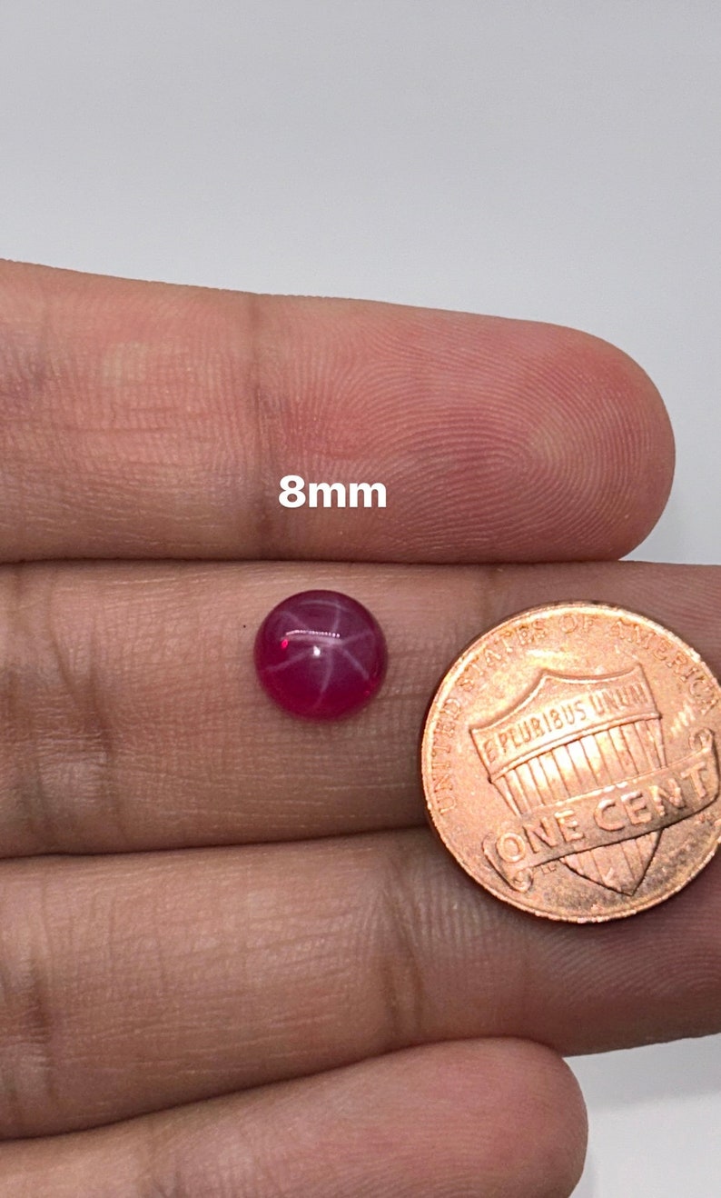 May include: A pink star ruby gemstone with a starburst pattern, measuring 8mm in diameter. The gemstone is held in a hand next to a US penny for size comparison.