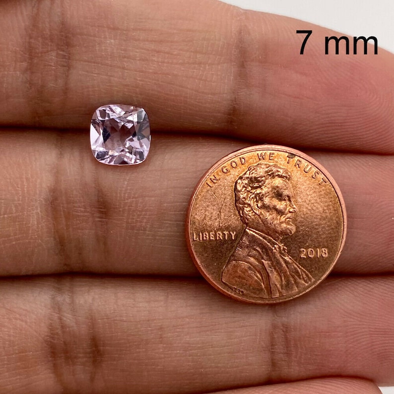 May include: A pale pink, cushion-cut gemstone measuring 7 mm in diameter, next to a US penny for size comparison.