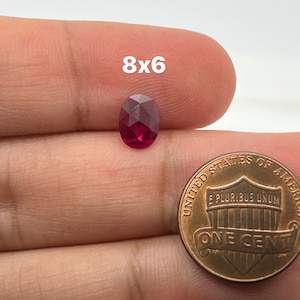 Puede incluir: Una piedra preciosa ovalada de color rojo intenso, de aproximadamente 8 mm por 6 mm, se sostiene en una mano junto a un centavo estadounidense para comparar el tamaño.