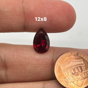 May include: A deep red pear-shaped gemstone, measuring 12x8, is shown next to a US penny for size comparison.