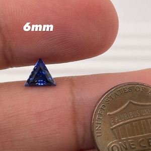 Puede incluir: Una piedra preciosa azul triangular de 6 mm en un dedo con una moneda de 1 céntimo de euro para comparar el tamaño.